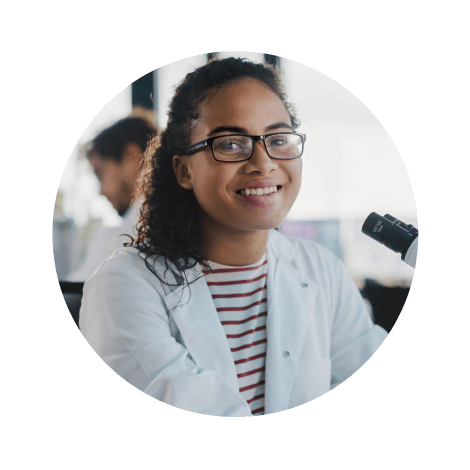 Joven interna con gafas sonriente con bata blanca de laboratorio sentada junto a un microscopio.